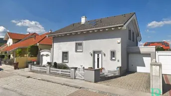 Graues Einfamilienhaus mit Vorgarten, Garage und gepflasterter Einfahrt unter blauem Himmel.
