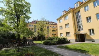 Gelbes Mehrfamilienhaus mit grüner Gartenanlage und Fahrradabstellplätzen unter blauem Himmel.
