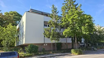 Mehrstöckiges Wohngebäude mit heller Fassade und umgebenden Bäumen unter blauem Himmel.