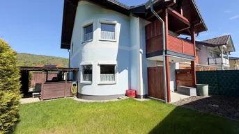 Hausfassade in hellem Blau mit Holzdetails, kleinem Garten und Carport an einem sonnigen Tag.