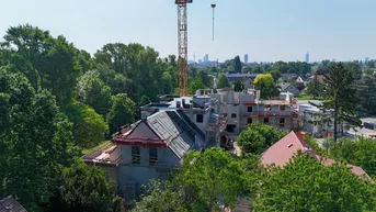 Luftaufnahme eines im Bau befindlichen Gebäudes mit Kran, umgeben von Bäumen und Stadtsilhouette im Hintergrund.