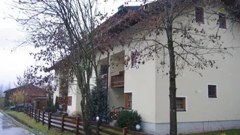 Mehrfamilienhaus mit traditioneller Architektur, umgeben von Bäumen und einem Holzzaun in ländlicher Umgebung.