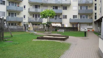 Gemeinschaftsgarten mit Sandkasten und Bänken vor einem Mehrfamilienhaus mit zahlreichen Balkonen.