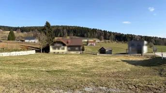 Weitläufige Grünfläche mit Blick auf bewaldete Hügel und vereinzelte Häuser in der Ferne unter blauem Himmel.