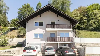 Einfamilienhaus mit Satteldach, Balkonen und Parkplätzen im Freien vor grüner Kulisse.