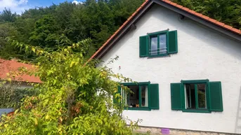 Weißes Haus mit grünen Fensterläden und rotem Ziegeldach vor bewaldetem Hügel.