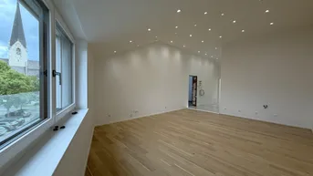 Helles, geräumiges Zimmer mit Holzparkettboden, vielen Deckenspots und Blick auf einen Kirchturm.