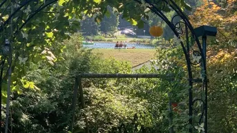 Idyllischer Gartenweg mit bewachsenem Torbogen und Blick auf einen See in der Ferne.