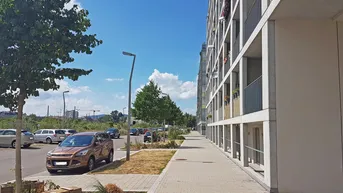 Moderne Wohngebäude mit Balkonen entlang eines Gehwegs und Parkplätzen unter blauem Himmel.