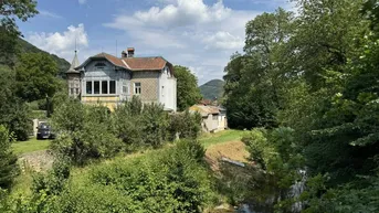 Historische Villa am Flussufer, umgeben von üppigem Grün und Bergen im Hintergrund.