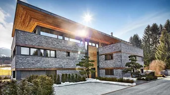 Modernes Gebäude mit Stein- und Holzfassade unter strahlendem Himmel in alpiner Landschaft.