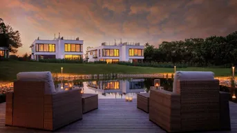 Moderne Häuser am Teich bei Sonnenuntergang mit gemütlicher Terrasse im Vordergrund.