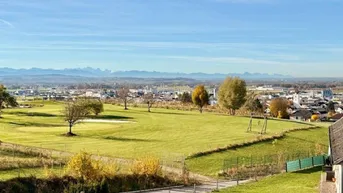 Weitläufiger Blick über eine grüne Landschaft mit Stadt und Alpenpanorama im Hintergrund.