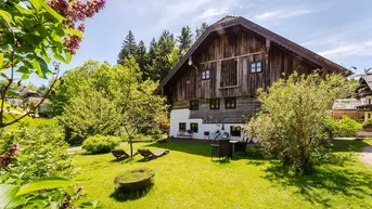 Charmantes Holzhaus mit großem Garten, Liegestühlen und blühenden Fliederbüschen unter blauem Himmel.