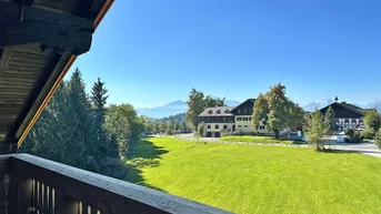 Blick vom Holzbalkon über eine weite grüne Wiese mit Gebäuden und Bergen im Hintergrund unter blauem Himmel.