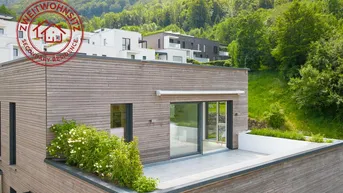 Moderne Architektur mit Holzfassade und großer Dachterrasse, umgeben von grünen Hügeln.