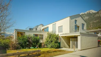 Modernes Gebäude mit heller Fassade, Gartenbereich und Bergblick unter blauem Himmel.