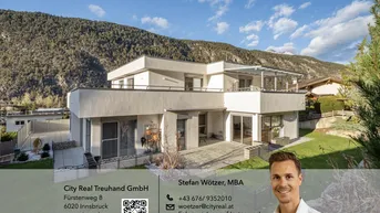 Moderne Villa mit mehreren Terrassen und Balkonen vor alpiner Bergkulisse und viel Grün.