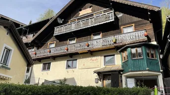Traditionelles Gebäude mit Holzfassade und mehreren Balkonen, umgeben von grünen Hecken und Bäumen.