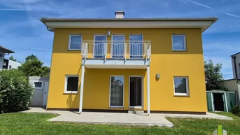 Gelbes Einfamilienhaus mit Balkon, kleiner Terrasse und grünem Garten unter blauem Himmel.