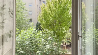 Blick aus einem Fenster in einen grünen Innenhof mit Bäumen und einem Wohngebäude im Hintergrund.