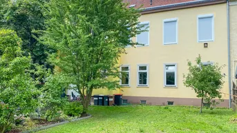 Rückseite des gelben Gebäudes mit Blick auf den grünen Garten und alte Bäume.