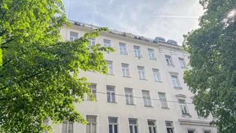 Helle Fassade eines Mehrfamilienhauses mit grünen Bäumen im Vordergrund unter bewölktem Himmel.