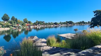 Weitläufiger Blick auf einen ruhigen See mit Häusern am Ufer unter blauem Himmel.