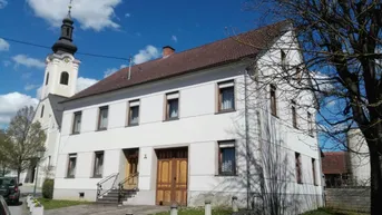 Weißes Wohnhaus mit braunem Ziegeldach und einer Kirche im Hintergrund unter blauem Himmel.