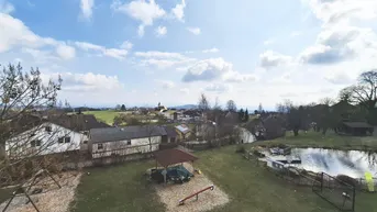 Weitläufiger Blick über eine ländliche Umgebung mit Häusern, einem Teich und einem Spielplatz unter blauem Himmel.