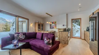 Heller offener Wohnbereich mit violetter Couch, Essbereich und Zugang zur Terrasse mit Bergblick.