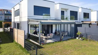 Ansicht des modernen Reihenhauses mit Garten, Terrasse und Balkonen, umgeben von einer grünen Landschaft.