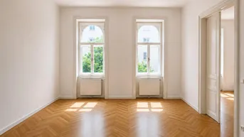 Helles Zimmer mit Fischgrätparkett, weißen Wänden und zwei großen Fenstern, die viel Licht hereinlassen.