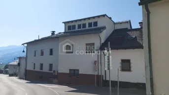 Gebäudeansicht von der Straße mit weißer Fassade und Blick auf die umliegende Berglandschaft.