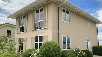 Modernes Einfamilienhaus mit beiger Fassade, weißen Fenstern und gepflegtem Vorgarten unter bewölktem Himmel.