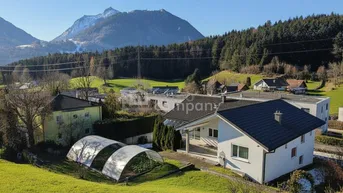 Haus mit Garten und Gewächshaus in ländlicher Umgebung mit Bergblick unter blauem Himmel.