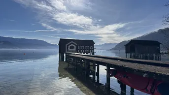 Idyllischer See mit traditionellen Bootshäusern und majestätischer Bergkulisse unter blauem Himmel.