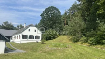 Weißes Landhaus mit dunklen Fensterläden, umgeben von einem weitläufigen Garten und Wald.