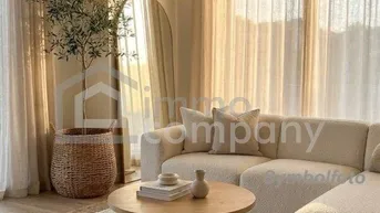 Helles und gemütliches Wohnzimmer mit beigem Sofa, Holzboden, großem Teppich und viel Tageslicht durch die Fenster.