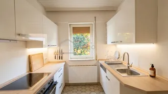 Helle Küche mit weißen Hochglanzschränken, Holz-Arbeitsplatten und einem Fenster mit Blick ins Grüne.