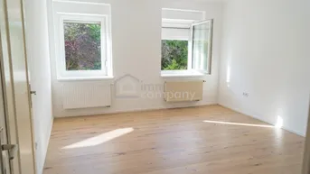 Heller, leerer Raum mit Holzboden, weißen Wänden und zwei Fenstern, die viel Tageslicht hereinlassen.