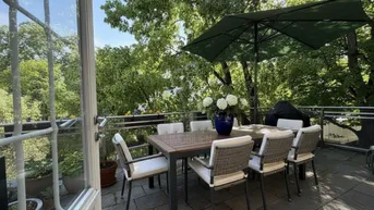 Sonnige Terrasse mit Esstisch, Stühlen und Sonnenschirm, umgeben von üppigem Grün.