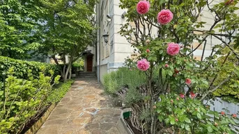 Steinweg führt durch einen blühenden Rosengarten zum Eingang eines eleganten Gebäudes.