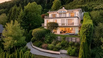 Elegante Villa am Hang mit mehreren Balkonen und üppiger grüner Umgebung bei Sonnenuntergang.