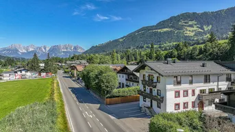Gebäudeansicht mit traditioneller Architektur, umgeben von grünen Wiesen und majestätischen Bergen unter blauem Himmel.