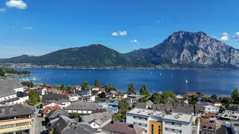 Panoramablick auf eine malerische Stadt am Seeufer, umgeben von majestätischen Bergen unter blauem Himmel.