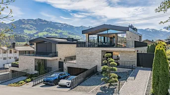Moderne Villa mit Steinfassade, großen Fenstern und Bergblick, umgeben von grüner Landschaft.