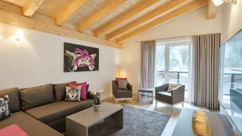 Gemütliches Wohnzimmer im alpinen Stil mit Holzbalkendecke, bequemer Couch und Zugang zum Balkon.