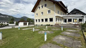 Gelbes Gebäude mit Satteldach und großem Grundstück, umgeben von Bergen unter bewölktem Himmel.