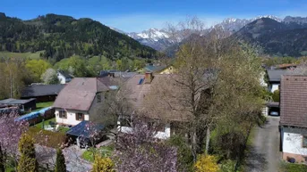 Luftaufnahme eines Wohngebiets mit traditionellen Häusern, umgeben von grünen Hügeln und majestätischen Bergen unter blauem Himmel.